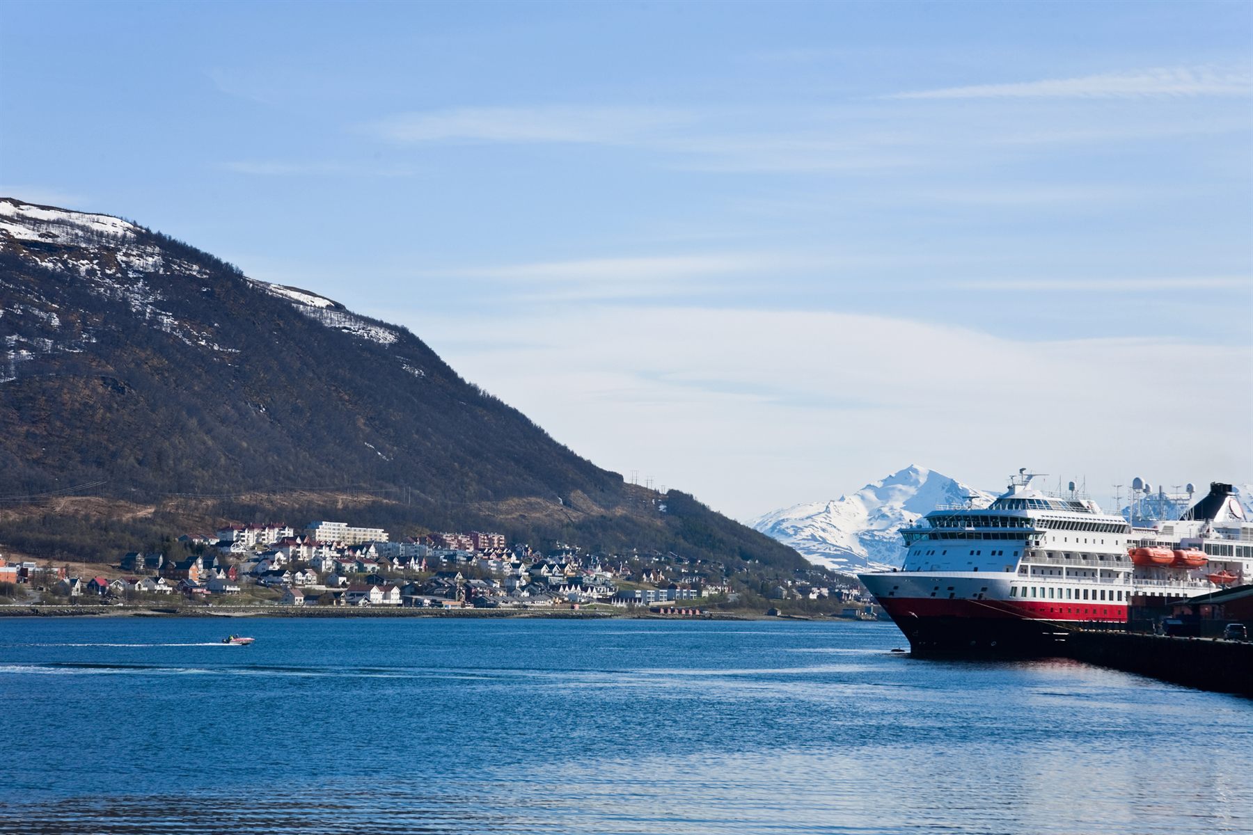 A red-and-black coastal ship sailing past a town below snowy mountains in northern Norway